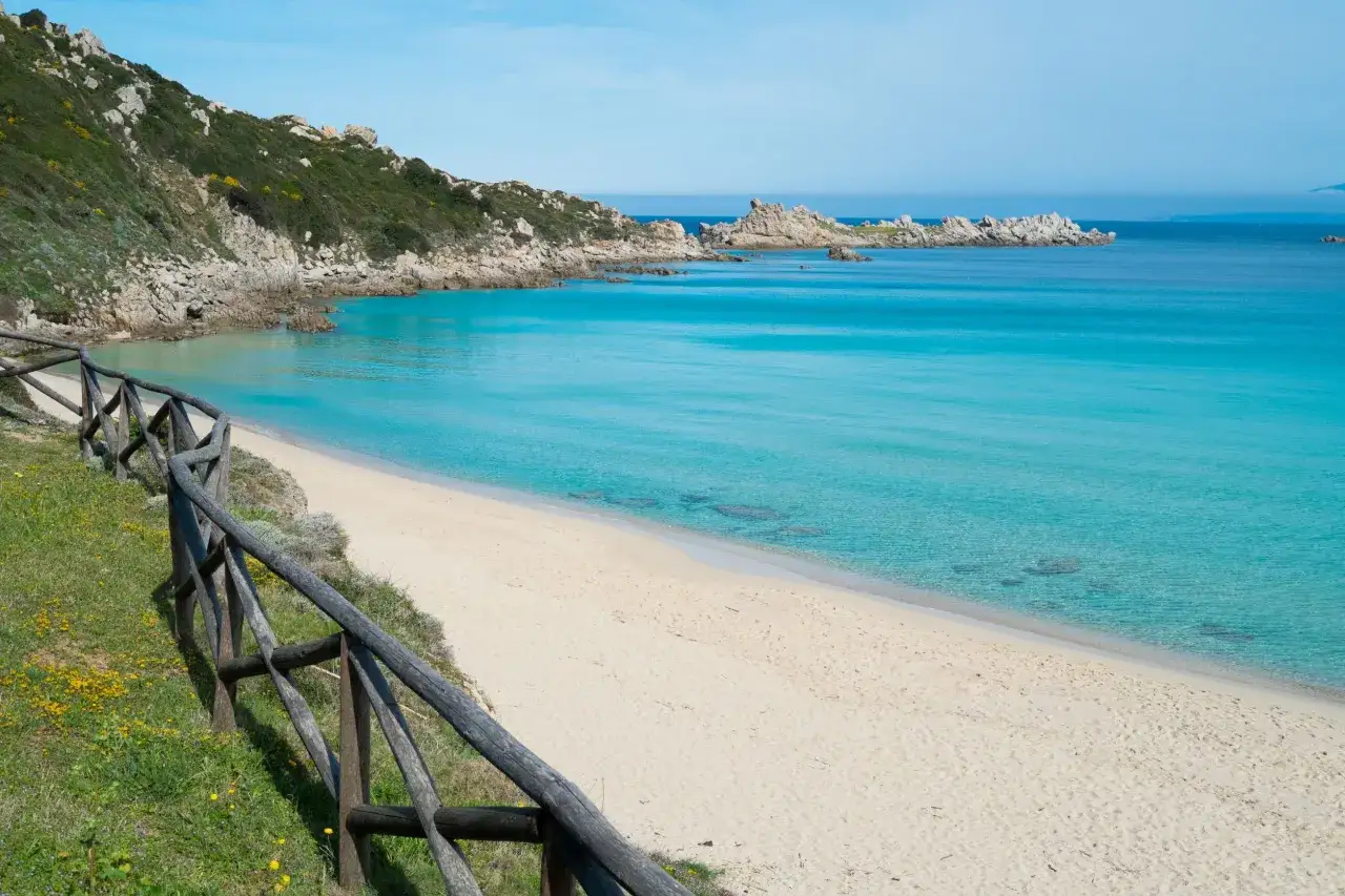 Spiaggia rena bianca con acque turchesi cristalline, rocce e vegetazione mediterranea. Una staccionata in legno delimita il sentiero erboso.