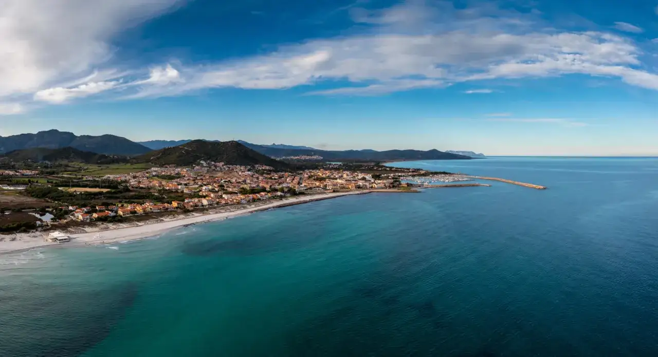 Vista aerea di Posada, Siniscola, con case colorate, spiaggia bianca e mare turchese sotto un cielo azzurro con nuvole bianche.