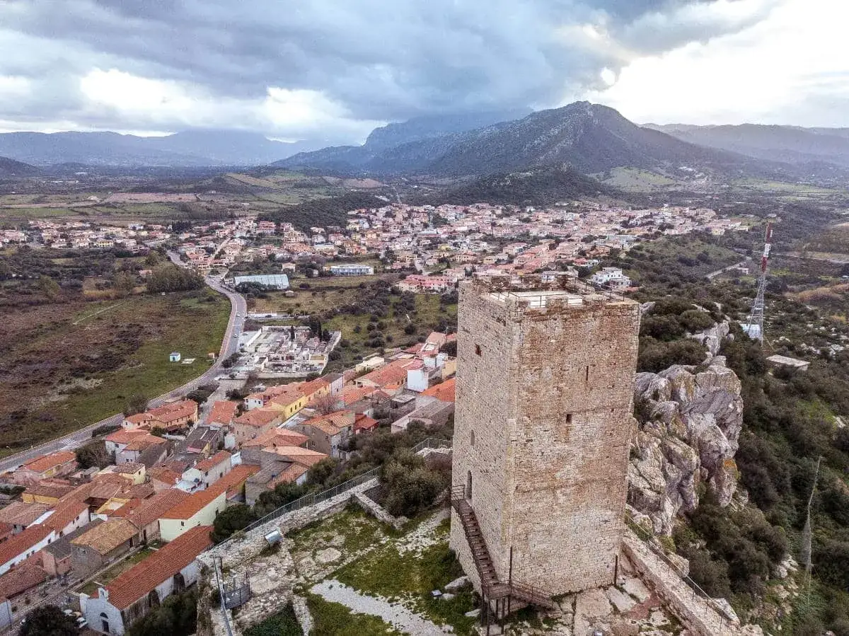 Torre medievale domina il borgo di Posada, in Sardegna, con montagne sullo sfondo.