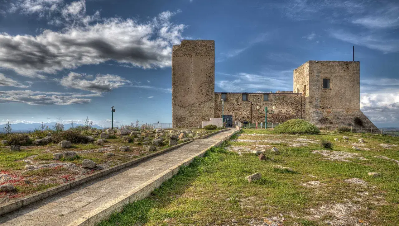 Sentiero lastricato verso il Castello San Michele, con torri in pietra e cielo azzurro con nuvole.