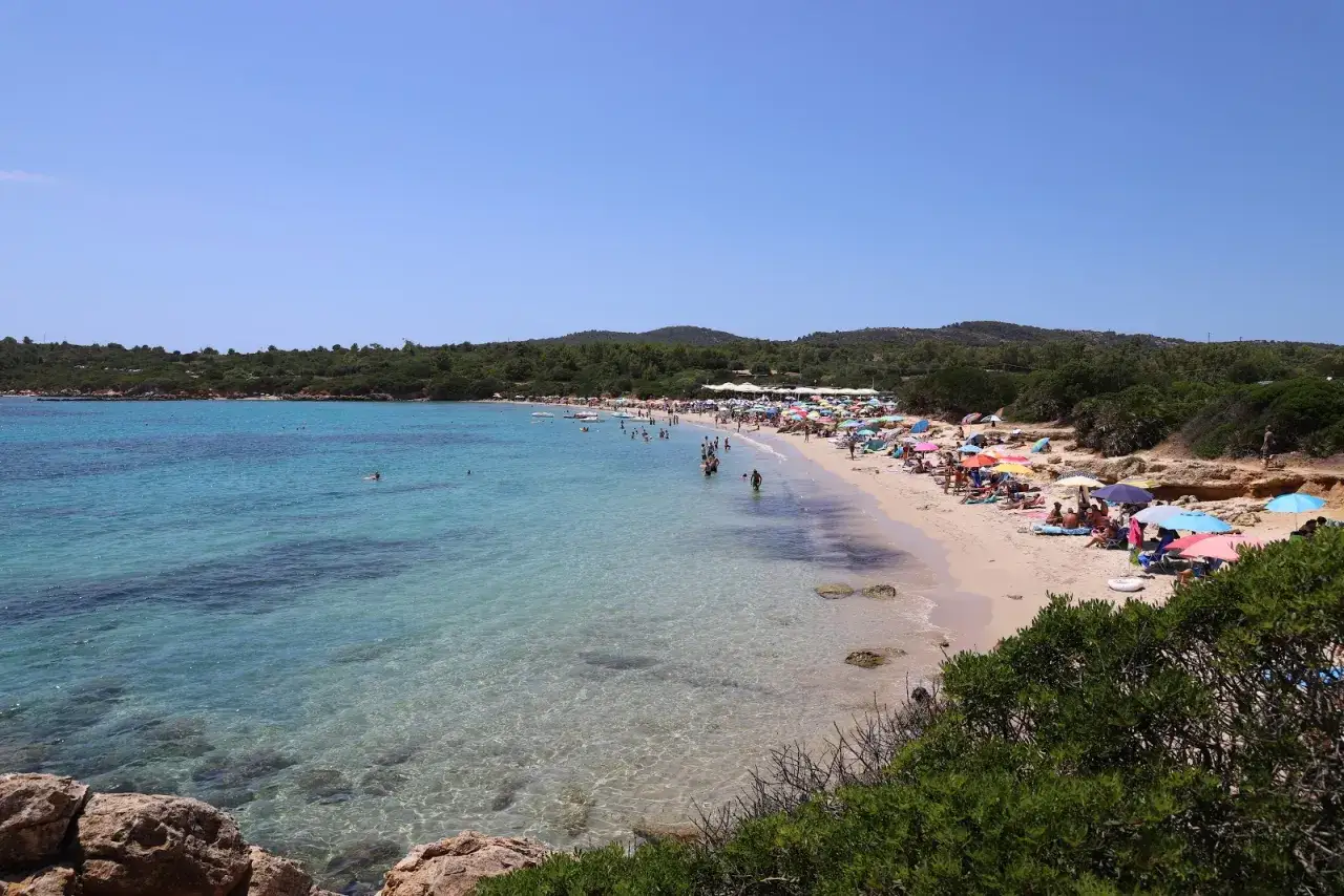 Una giornata di sole alla spiaggia Lazzaretto di Alghero, con acque cristalline e tanta gente che si gode il mare sotto ombrelloni colorati.