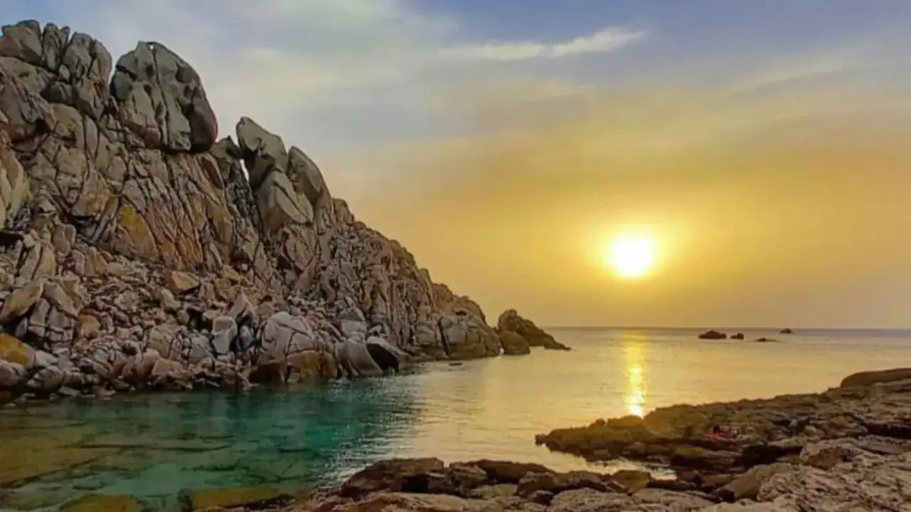 Scogliere rocciose bagnate da acque cristalline, con il sole che tramonta sulla valle della luna, creando riflessi dorati sul mare.