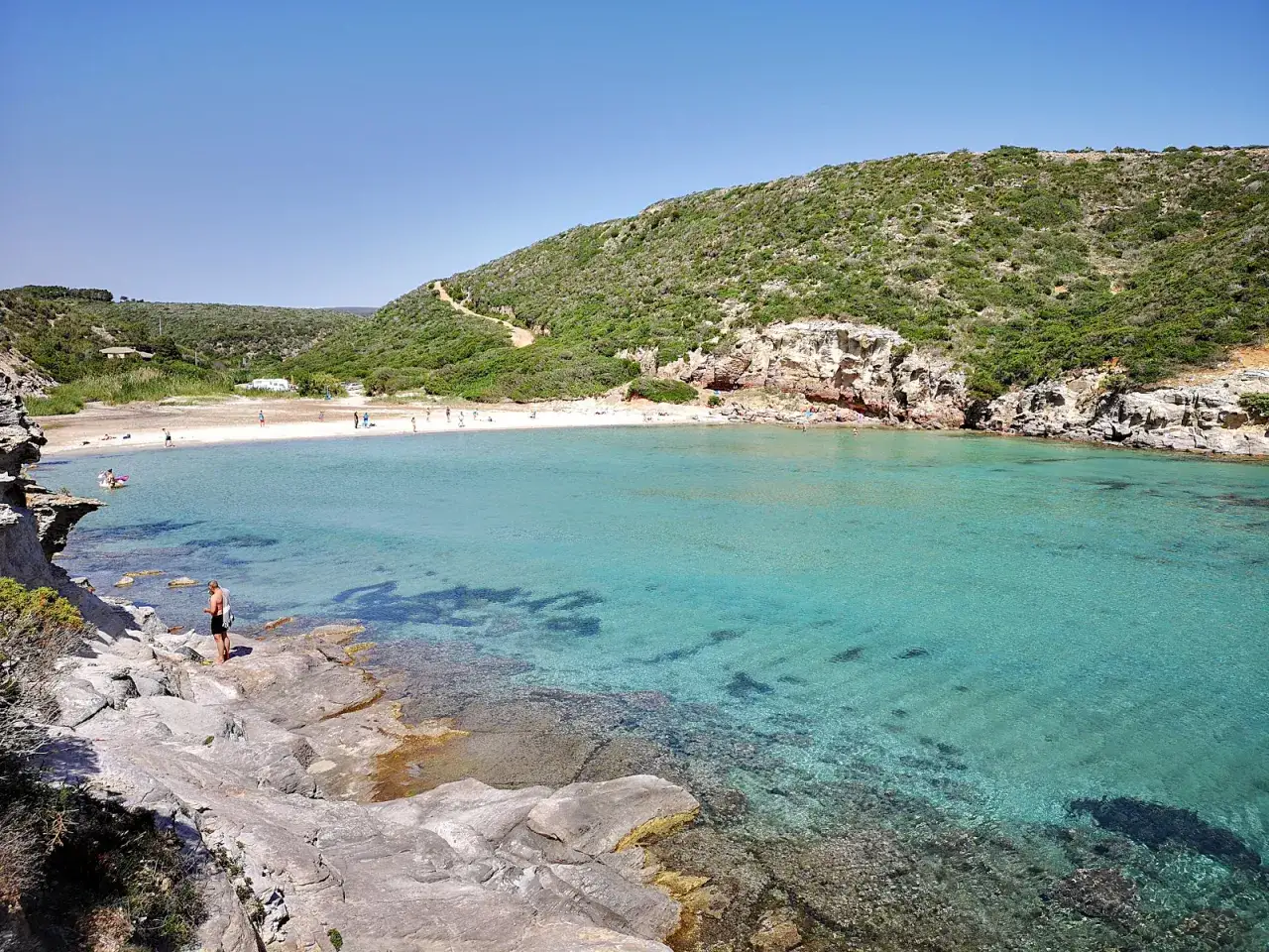 Uomo su rocce guarda la splendida spiaggia cala lunga con acque turchesi e collina verdeggiante.