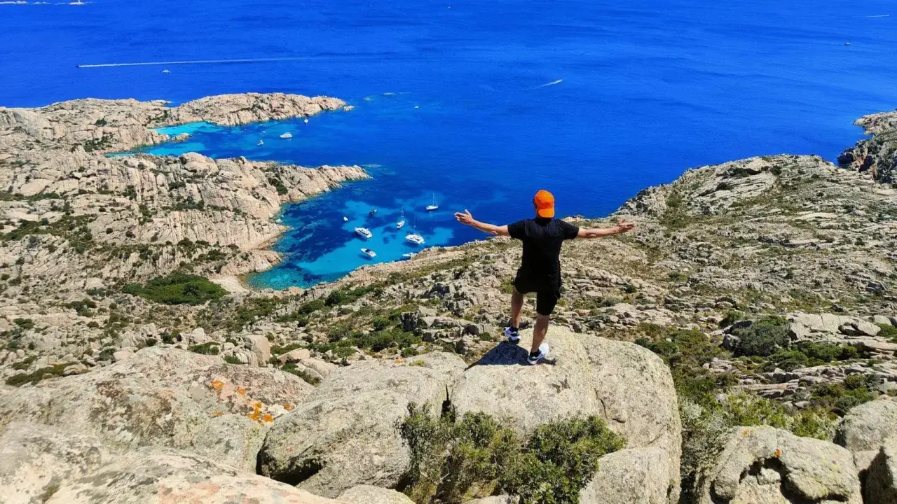 Uomo con cappellino arancione ammira la spiaggia di Cala Coticcio, con le sue acque cristalline e le barche all'ancora.
