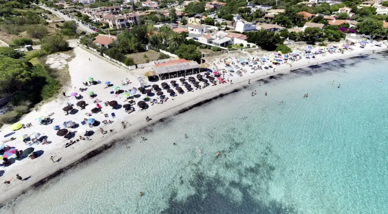 Vista aerea della spiaggia del pellicano, con ombrelloni colorati, bagnanti e acque cristalline. Case e vegetazione sullo sfondo.