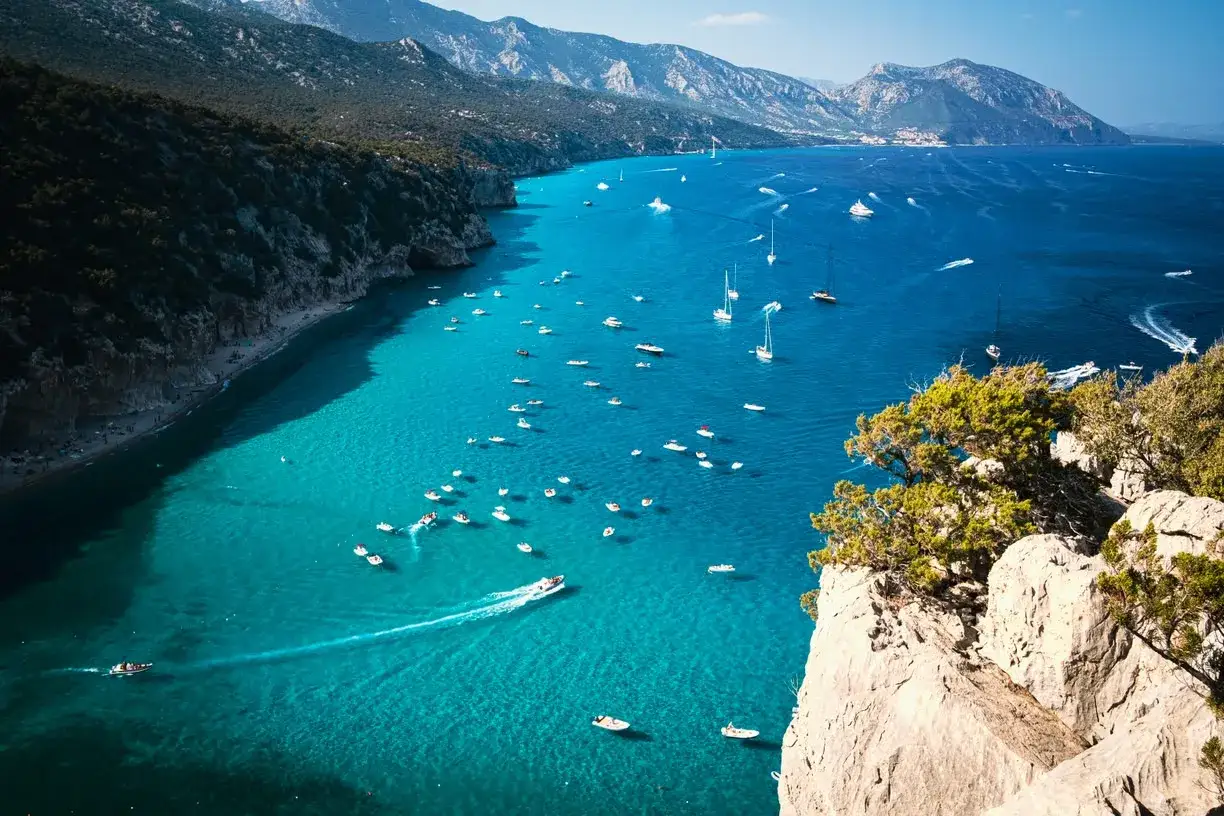 Un tratto di costa con acque cristalline e montagne sullo sfondo, un vero gioiello tra le spiagge più belle in Sardegna, punteggiato da numerose imbarcazioni.