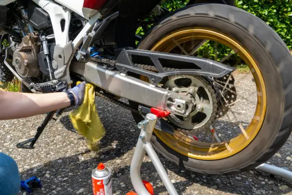 Mano con guanto pulisce catena moto con panno giallo. Spray pulitore catena e spazzola a terra.