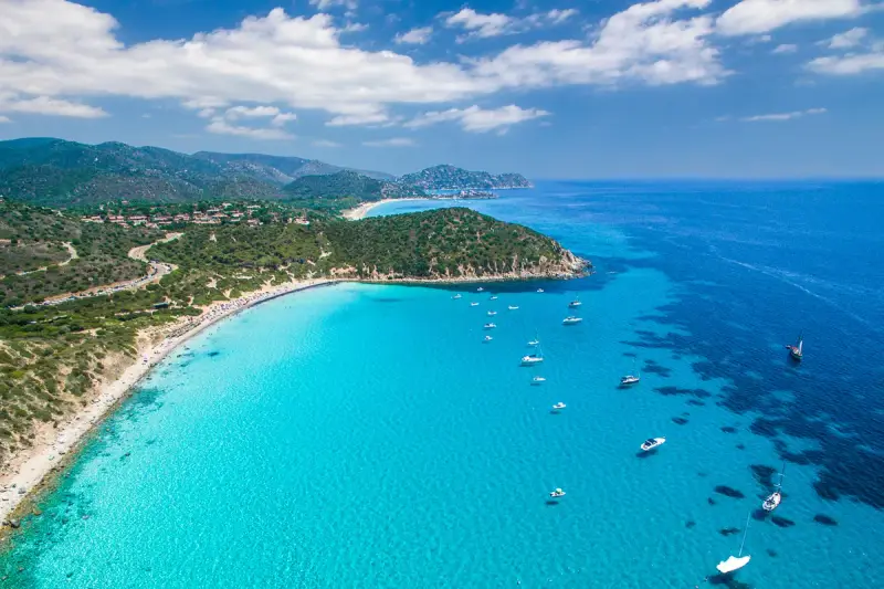 Spiaggia di Chia, Sardegna: acque cristalline che sembrano mari pintau, con barche a vela e un litorale affollato.