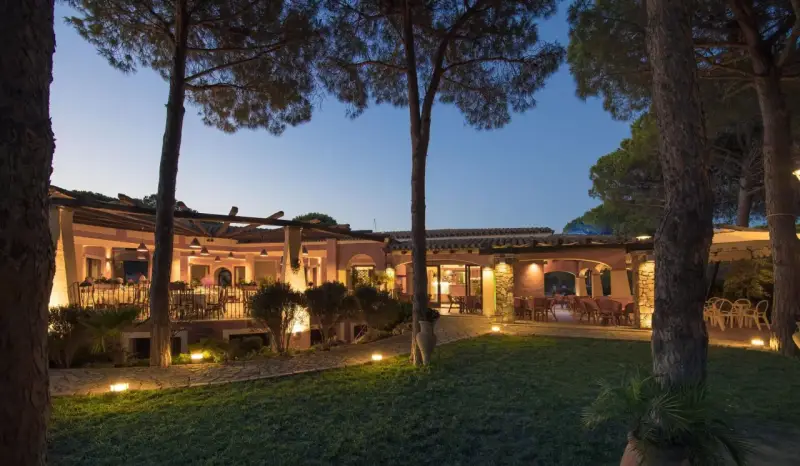 La sorgente Villasimius: un ristorante con tavoli all'aperto illuminati da luci soffuse, circondato da pini e un prato curato.