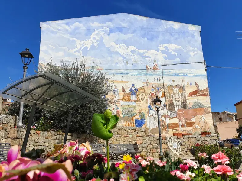 Un murale colorato a Loceri, Sardegna, raffigura scene di vita marina e rurale con persone, carri e animali sotto un cielo azzurro e nuvoloso.