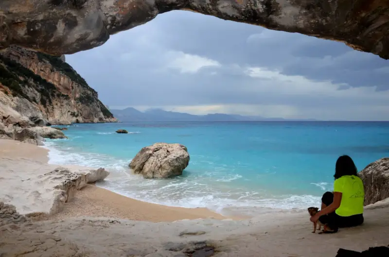 Vista da una grotta su Cala Goloritzé, con mare turchese, scogliere e una donna con un cane.