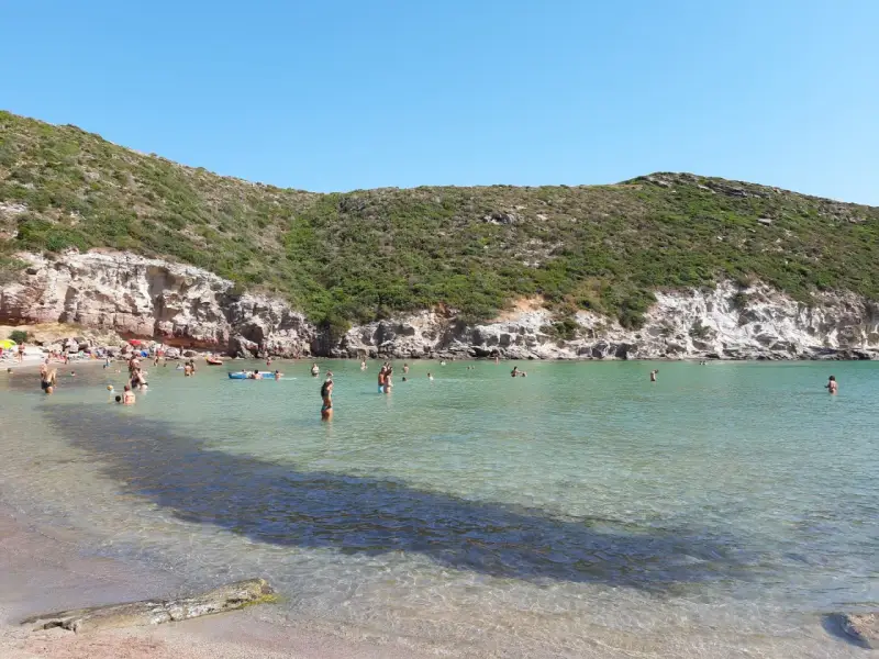 Persone si godono il sole e il mare cristallino nella spiaggia cala lunga, con colline verdi e rocce a fare da cornice.