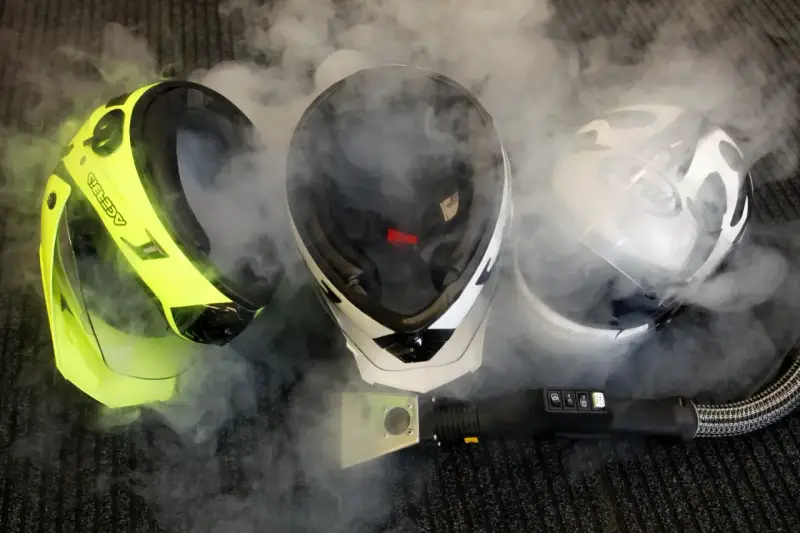 Tre caschi, uno giallo fluo, uno bianco e nero, e uno bianco, sono avvolti dal vapore. Un accessorio per la pulizia a vapore è posizionato davanti a loro, suggerendo che il casco non deve essere pulito con solventi.