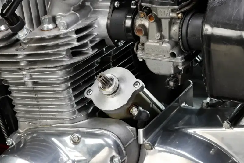 Dettaglio del motore di una moto: il test del motorino di avviamento è in primo piano, con il carburatore e le alette di raffreddamento visibili.